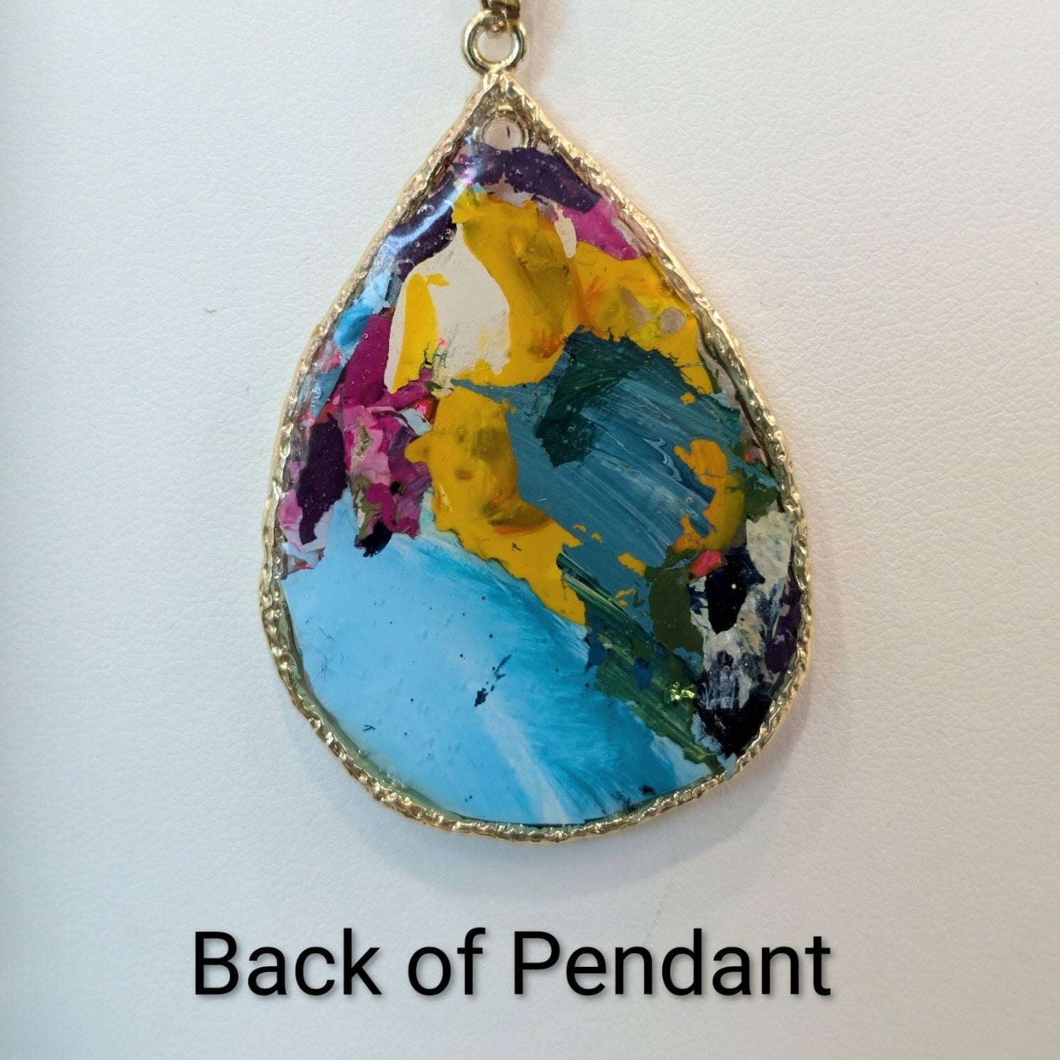 Two side-by-side product views of a teardrop pendant necklace on a white background, labeled 'Front of Pendant' and 'Back of Pendant,' against a dark gray surrounding backdrop. The pendant has a gold-tone chain and border with a colorful abstract floral design in blue, yellow, purple, pink, and white.